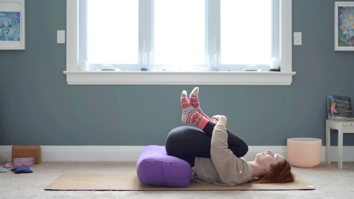 Yoga with Kassandra in a reclined supported bridge with both knees drawn into her chest