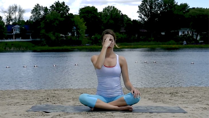 Yoga With Kassandra sits on a yoga mat in a cross-legged position practicing a breathwork technique for yoga for tension headaches.