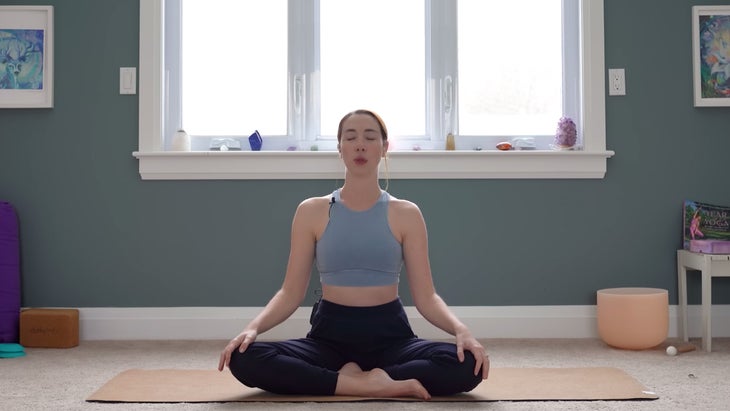 Yoga With Kassandra sits in a cross-legged position with her hands on her knees and her eyes closed while practicing yoga for stress relief.