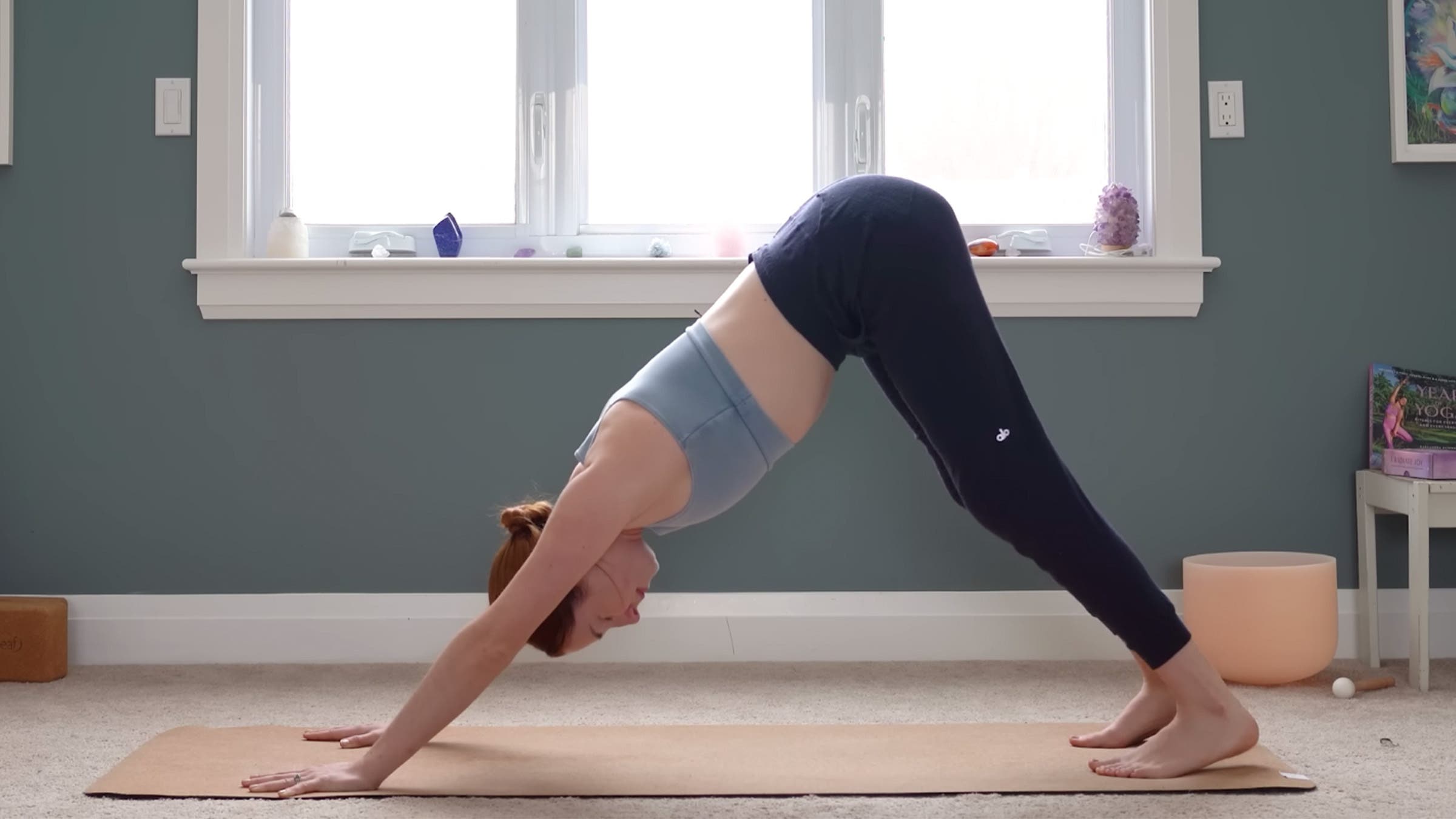 Yoga With Kassandra practicing Downward-Facing Dog with her hand and feet on the mat, shoulder-distance apart, and her hips lifted toward the ceiling in an inverted V-shape with her body while practicing yoga for stress relief.