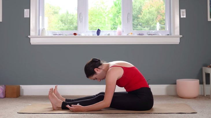 Woman sitting on a yoga mat with her legs in front of her as she slowly bends forward