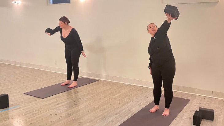 Women on yoga mats balancing blocks on their hands as they make circles around their heads