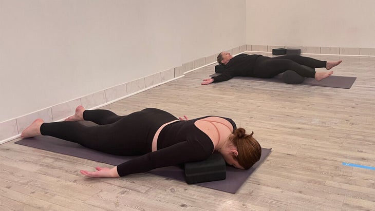 Woman lying prone on a yoga mat with blocks beneath her shoulders.