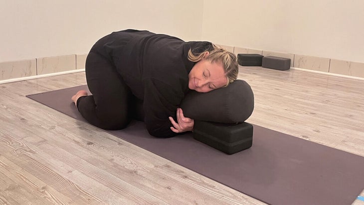 Woman in Child's Pose with support beneath her chest in the form of a bolster resting on a block