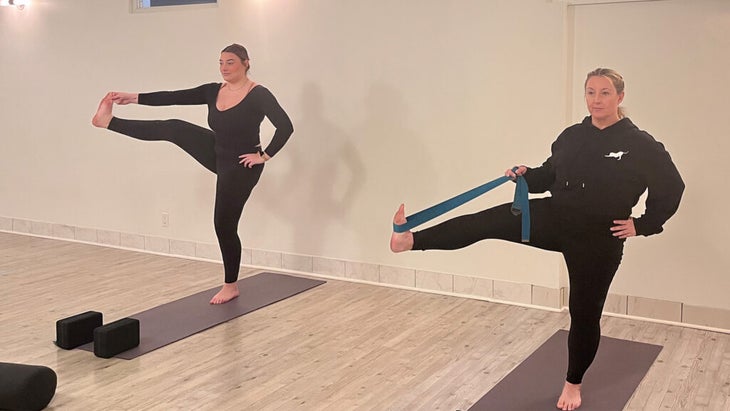 Students standing on mats practicing a pose with yoga props, including a strap.