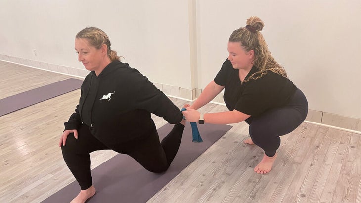 A yoga student on the mat and a teacher putting a yoga prop, a strap, in the student's hand
