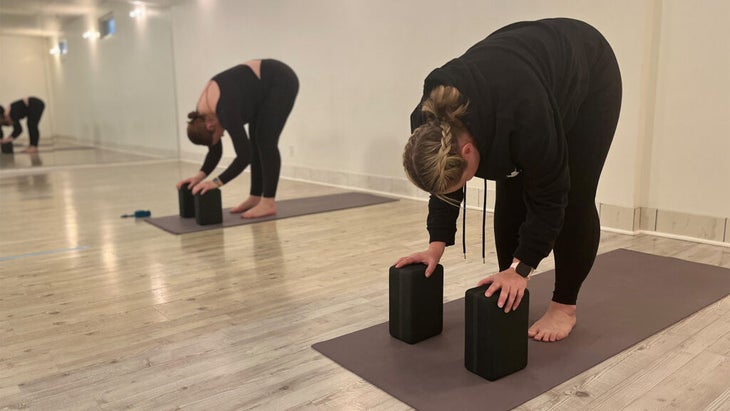 Yoga students on a mat with yoga props or blocks beneath their hands while bending forward