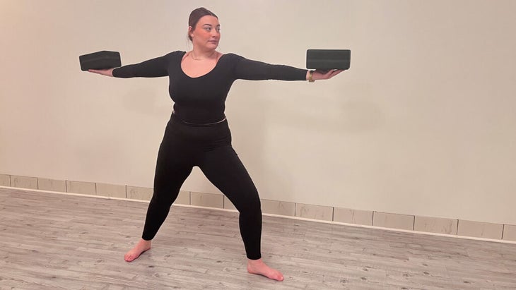 Woman on a yoga mat in Warrior 2 pose balancing blocks on her outstretched arms as a strengthening pose