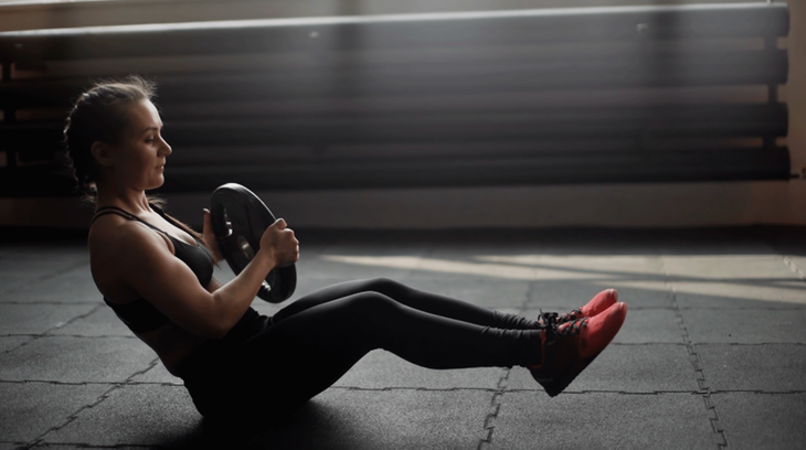 A woman sits on a yoga mat and practices a Russian twist by raising her legs and twisting her trunk from side to side.