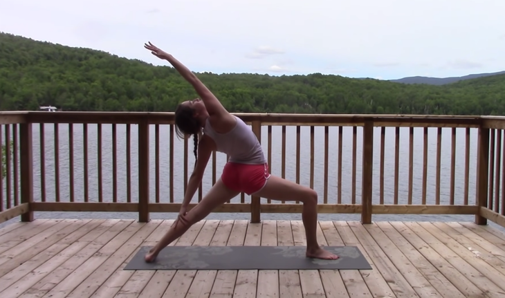 Yoga With Kassandra seated cross-legged outside on a mat practicing a side stretch during reverse warrior pose.