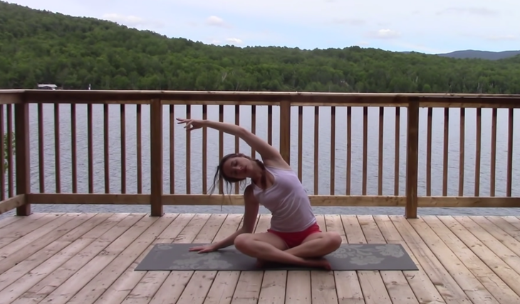 Yoga With Kassandra practices a seated side bend with her legs crossed and her left arm alongside her ear in a 20-minute yoga video