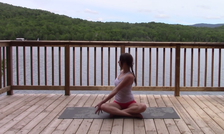 Yoga With Kassandra seated cross-legged outside on a mat practicing a side stretch and twist during a 20 minute yoga class