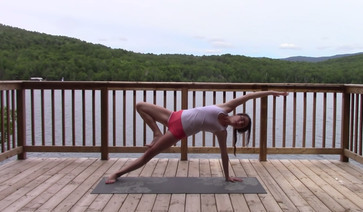 Yoga With Kassandra seated cross-legged outside on a mat practicing a side stretch in Side Plank during a 20 minute yoga class