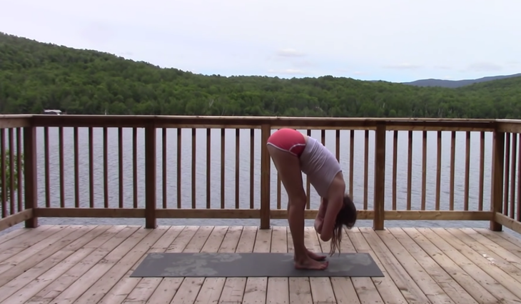 Yoga With Kassandra seated cross-legged outside on a mat practicing a standing forward bend during a 20 minute yoga class