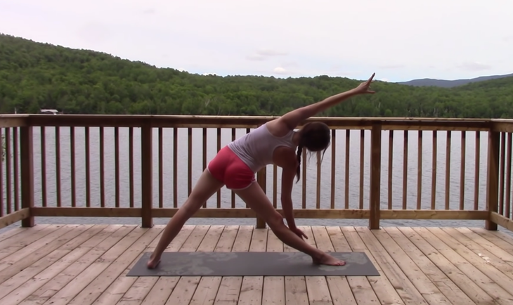 Yoga With Kassandra seated cross-legged outside on a mat practicing a side stretch during Triangle Pose.