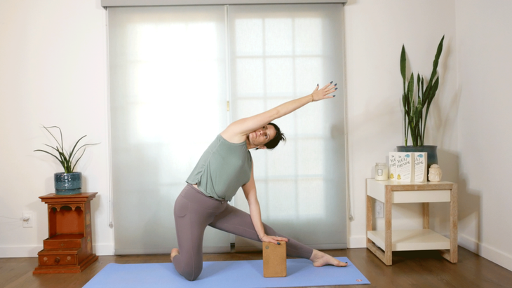 A yoga teacher practices Gate Pose by leaning her left hand on a block and reaching her right arm alongside her ear.