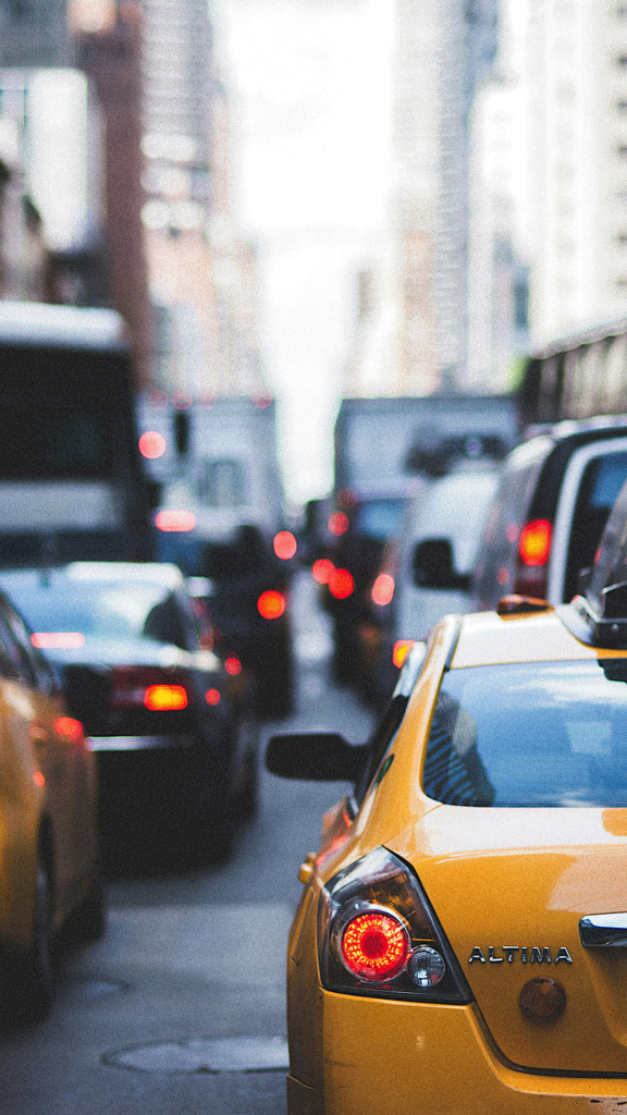 Traffic backed up on a narrow city street with the taillights of taxis and other cars beneath tall city buildings.