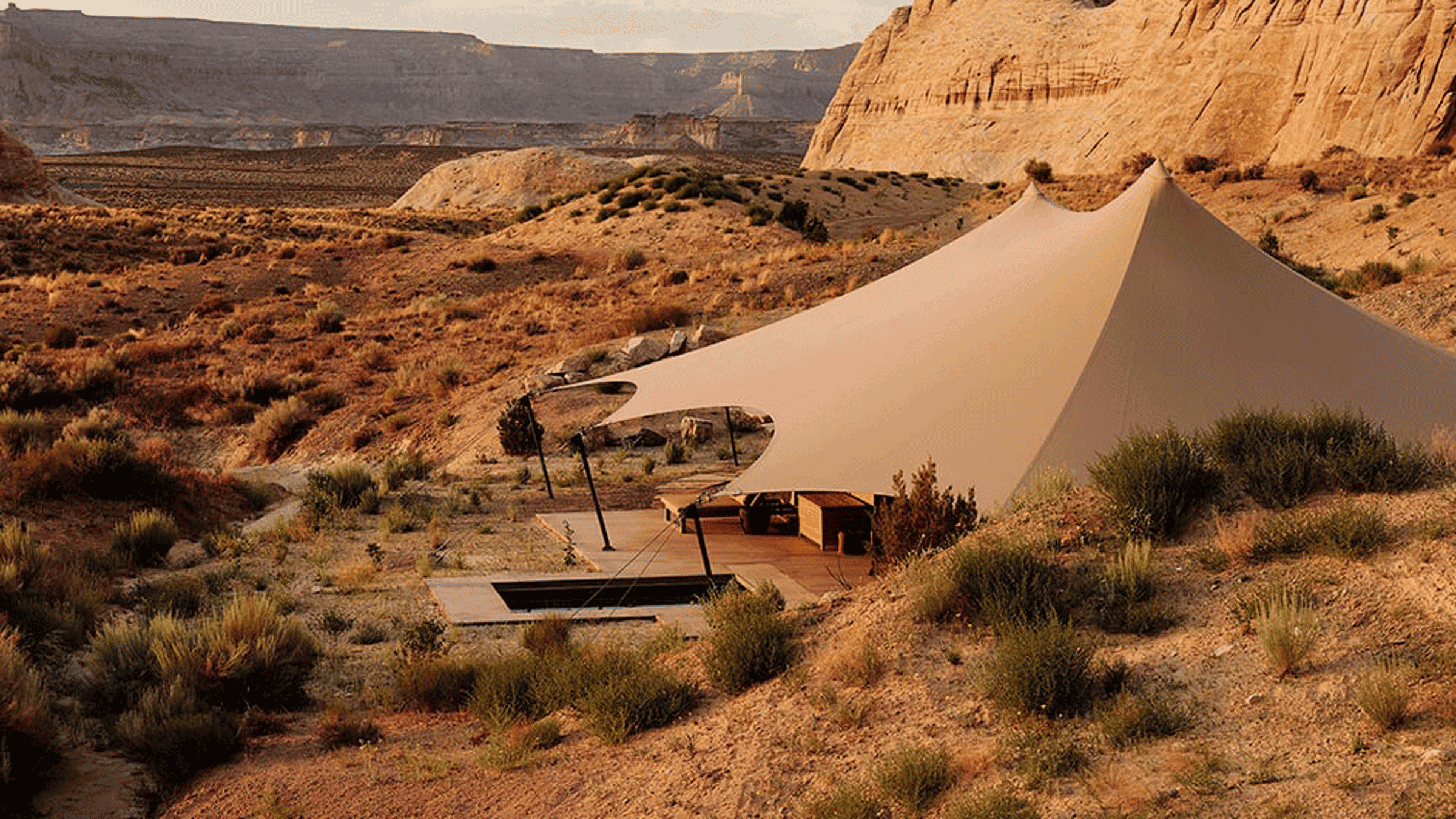 A desert landscape with a beige tent pitched on the sand among sparse shrubs.