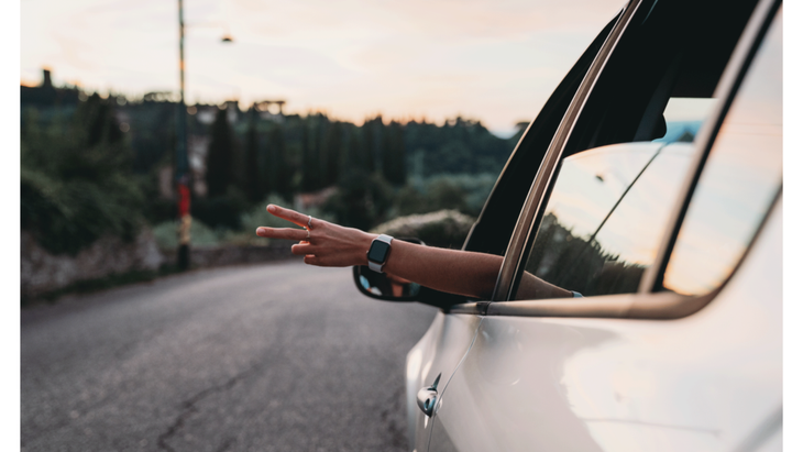 Young woman driving a car and throwing peace fingers out the driver side window to the car behind her