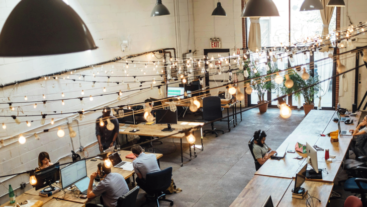 An overhead shot of a cool co-working space in Brooklyn with remote workers and their laptops sitting at long tables with strings of lights from the ceiling.