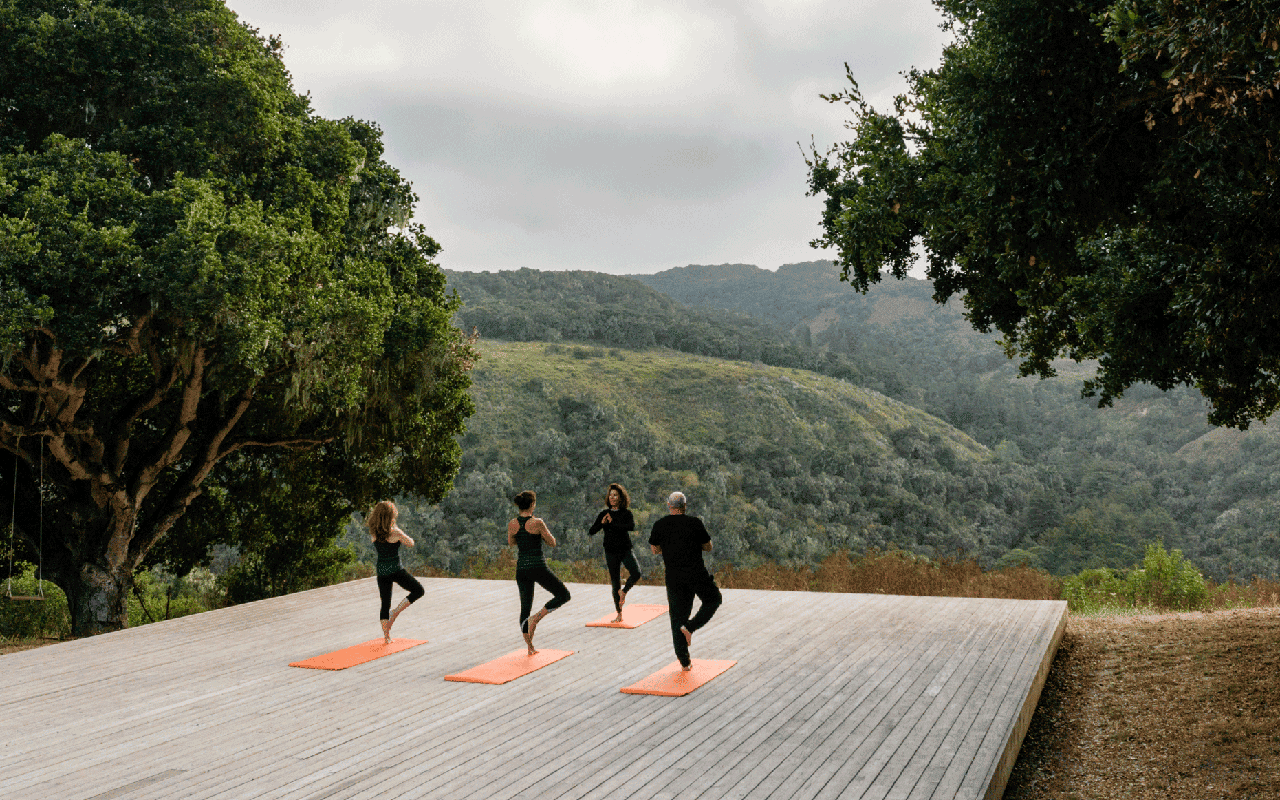 Three people doing yoga on a wooden deck overlooking forest and mountains.