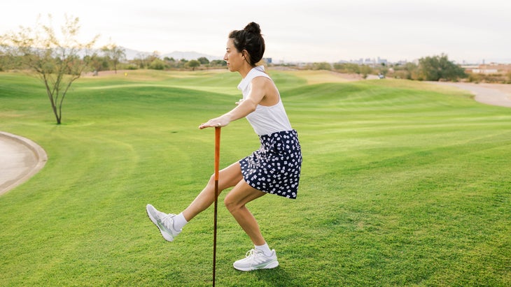 A woman performs yoga poses on a golf course with a club, doing a single leg squat on a gold course.