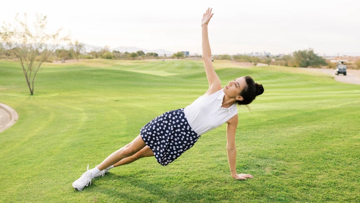 A woman performs yoga poses on a golf course with a club, in a plant pose with her arm raised to her side