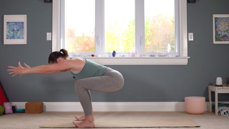woman on a yoga mat practicing a variation of Chair Pose in which she bends the knees deeply and reaches her arms straight forward