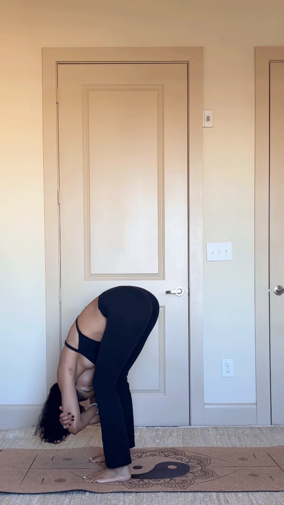 Woman standing in a forward bend at the top of her yoga mat while holding opposite elbows and swaying side to side