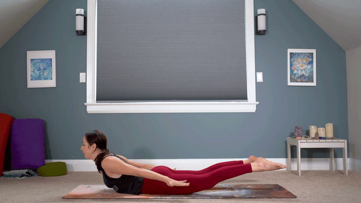 Yoga With Kassandra practicing Locust Pose on a yoga mat with her arms alongside her legs and her feet and chest lifted off the mat