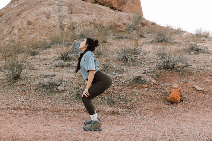 Woman hiker on a trail practicing a standing hiking stretch by bending her knees and arching her back