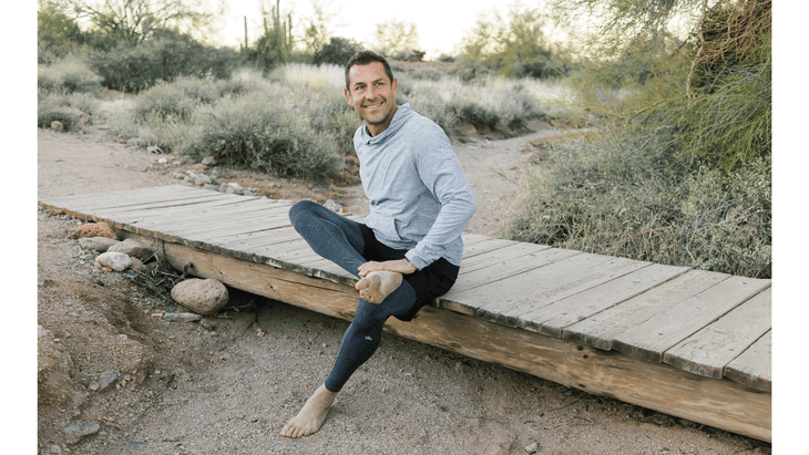 Man doing a figure 4 hiking stretch while sitting on a small bridge during a break on the trail.