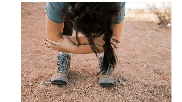 Woman stretching her hamstrings on a hiking trail by folding forward and grabbing opposite elbows.