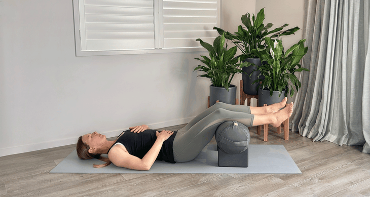 Woman lying on a yoga mat with her calves resting on a bolster placed on blocks.