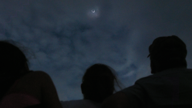 The solar eclipse with the moon blocking the sun in a dark sky with the silhouette of people watching below