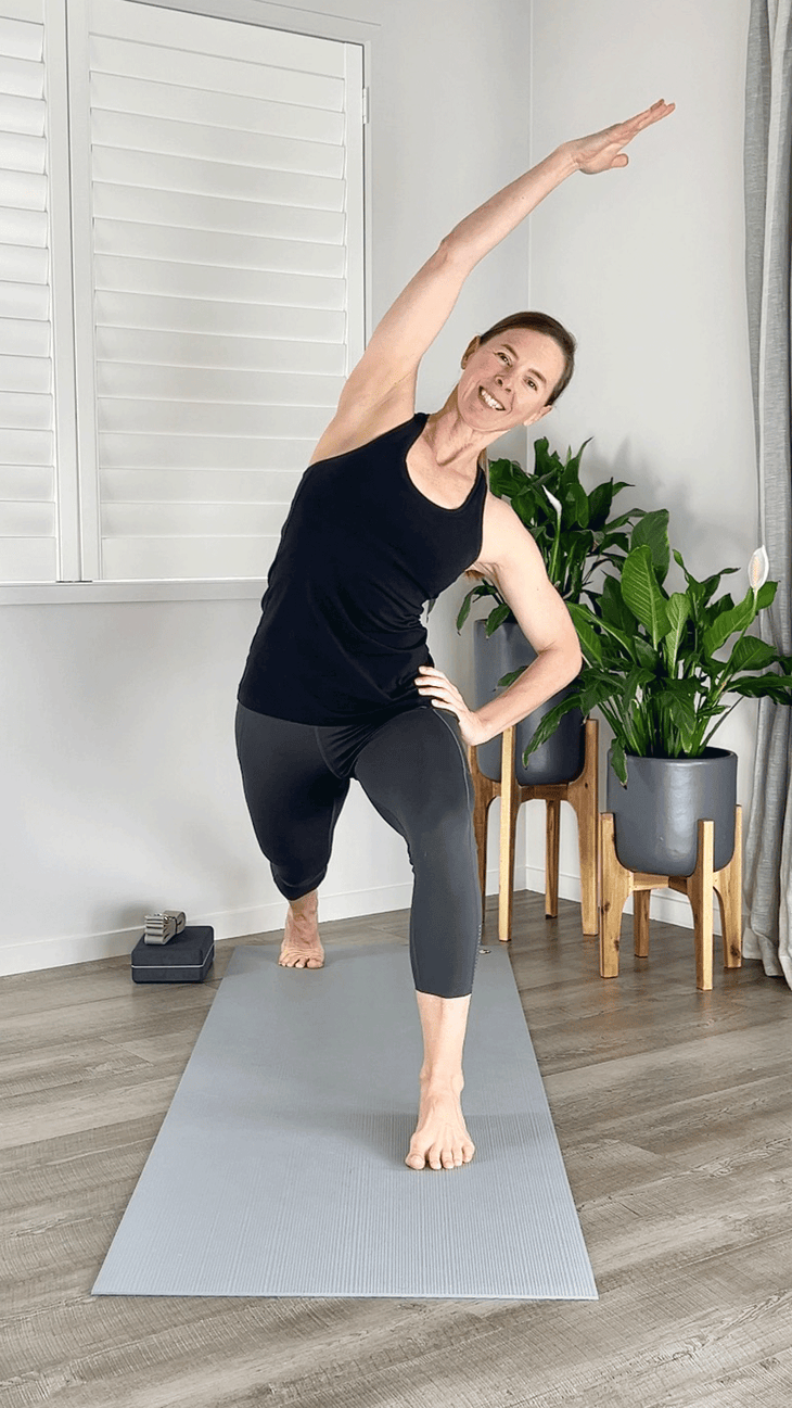 Woman on a yoga mat taking a side bend by reaching one arm overhead and leaning to the side