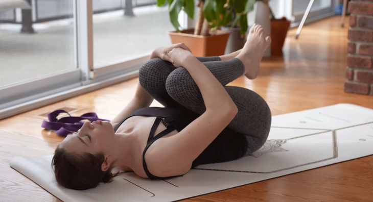 Woman lying on her back on a yoga mat practicing stretches for sleep by hugging her knees toward her chest