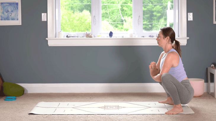 Woman in a squat at the back of a yoga mat with her hands in anjali mudra at her chest