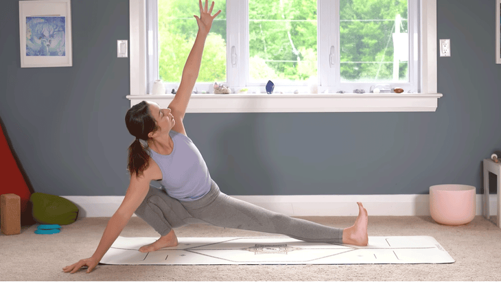 Woman in khaki green yoga pants and lavendar sports bra in a Side Lunge (Skandasana) on a yoga mat