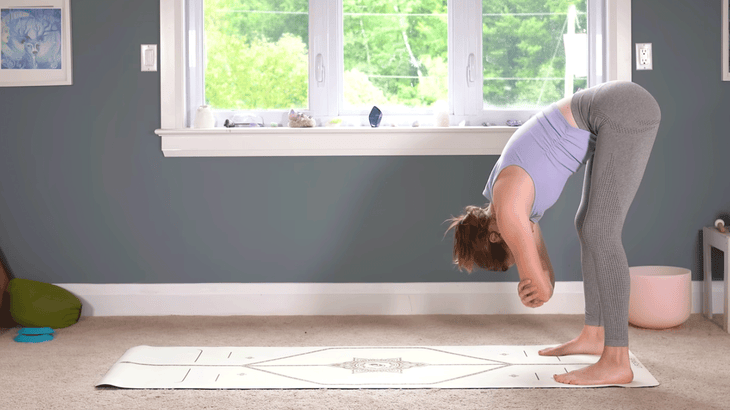 Woman standing at the back of a yoga mat in a forward bend with her hands on opposite elbows