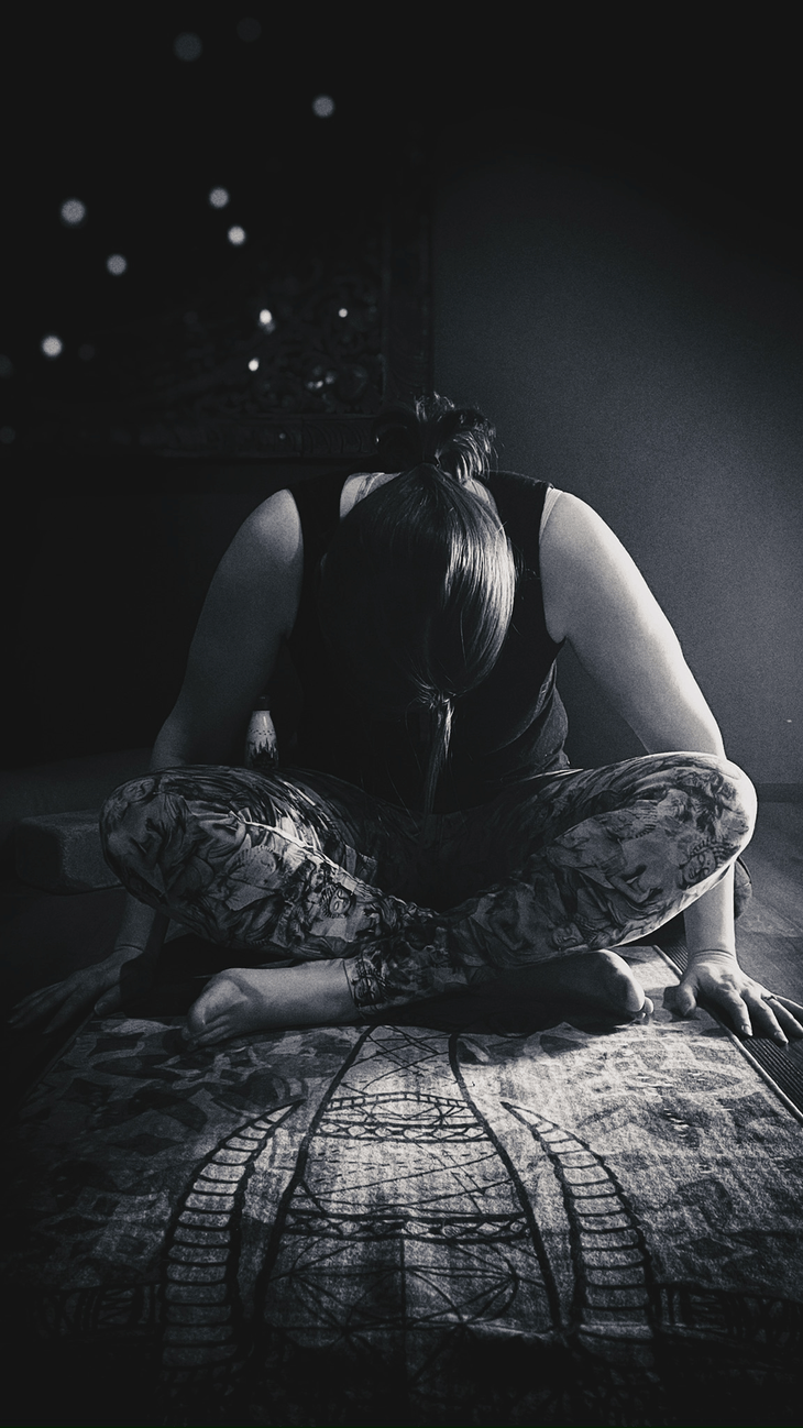 Black and white photo of the yin yoga pose named broken shoelace with a woman sitting cross-legged with knees over ankles while leaning forward.