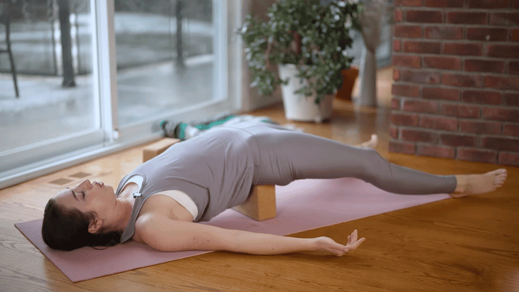 Woman lying on a yoga mat with a block beneath her hips in Supported Bridge