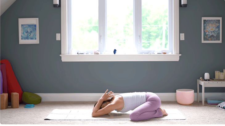 Woman in Child's Pose or Balasana by kneeling on a yoga mat with her chest close to the floor and her palms together at the base of her neck