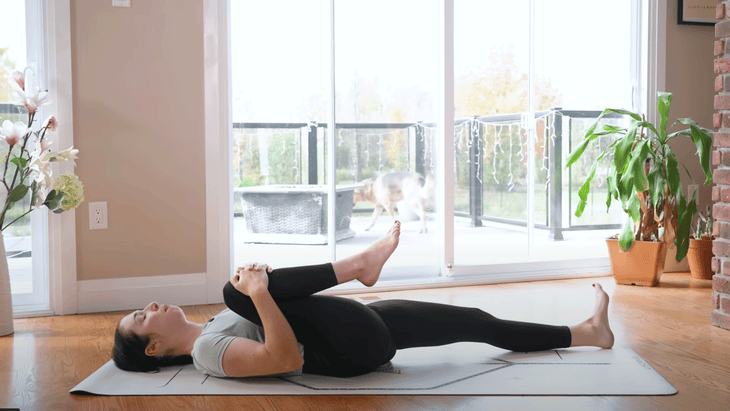 Woman lying on a yoga mat during a 15-minute morning yoga practice as she keeps her left leg straight and draws her right thigh into her chest