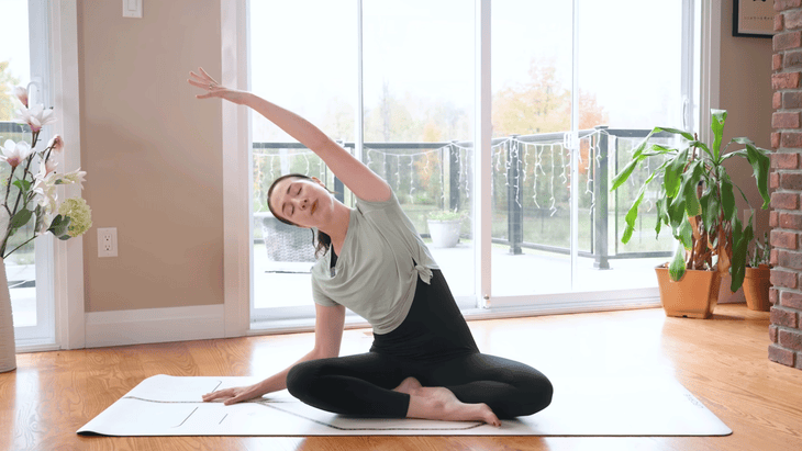 Woman sitting on a yoga mat practicing 15-minute morning yoga with a seated side stretch