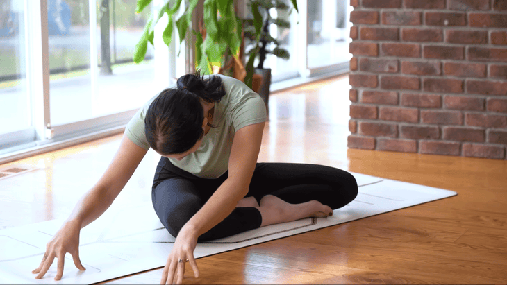 Woman sitting cross-legged on her yoga mat walking her hands to the side and folding forward 