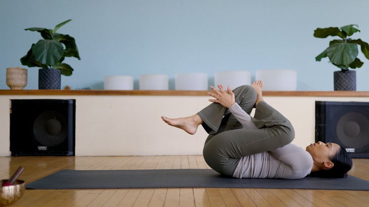 Woman reclined on a yoga mat with one knee drawn toward the chest in a Reclined Figure 4