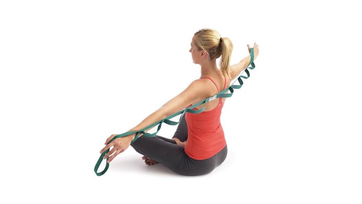 Image of woman sitting down and gripping a green Stretch Out Strap behind her back with both arms extended.