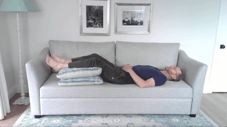 Man lying on a couch with cushions beneath his legs in a version of Legs Up the Wall