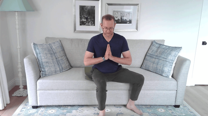 Man sitting on the edge of a couch with his hands at his chest in a version of Tree Pose